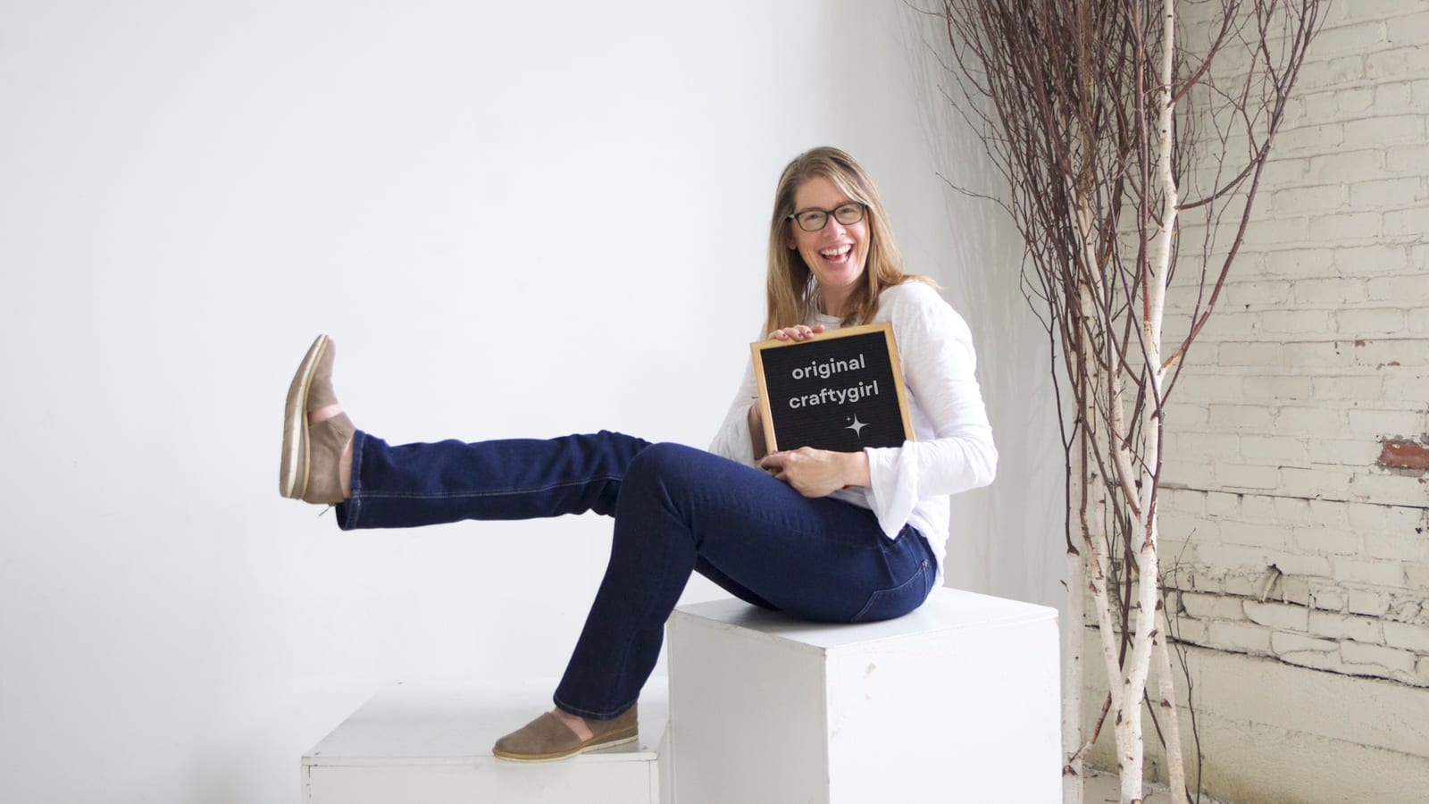 The founder of craftygirl sitting on a white plinth, laughing, holding a letterboard that reads 'original craftygirl'
