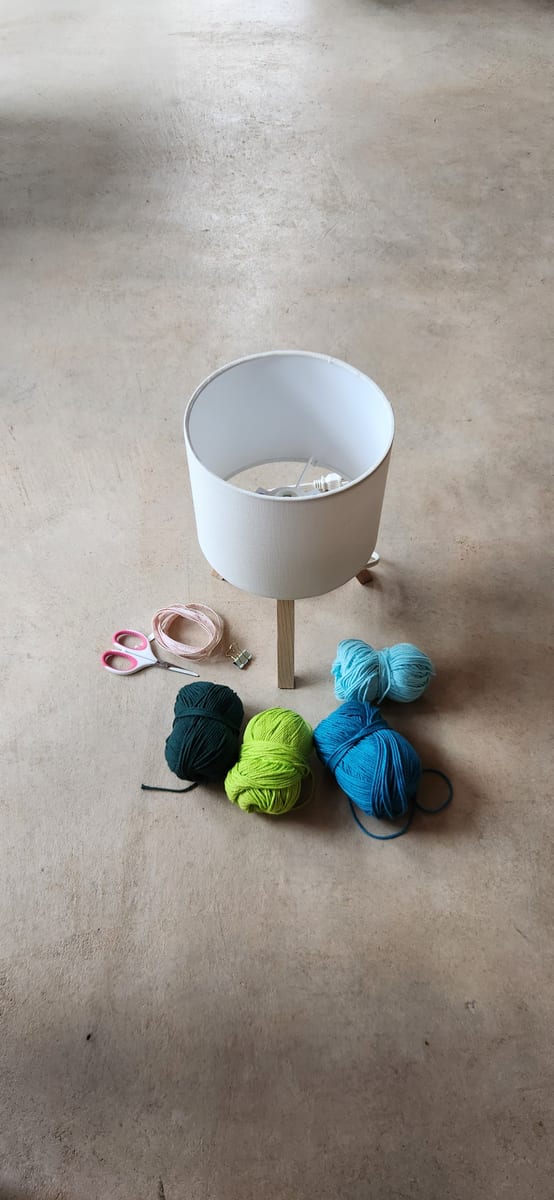 A bare white lampshade on a wooden base beside four balls of yarn